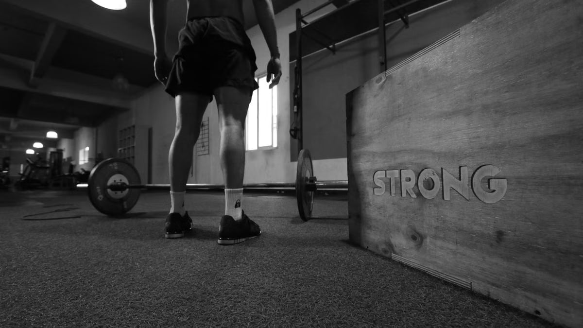 Black and white photo of a man in a gym with barbell and 'Strong' box.